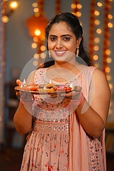 Vertical portrait shot of Happy Indian young woman looking at camera by holding diwali diya lamp at home - concept of