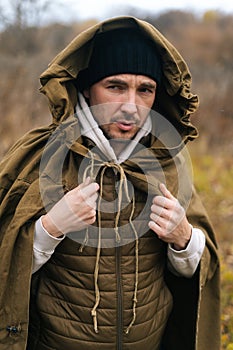 Vertical portrait of skilled survivalist male wearing green raincoat tent standing in thicket of bushes in cold overcast photo