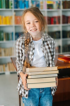 Vertical portrait of smiling elementary child school girl holding stack of books in library at school, looking at camera