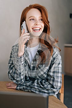 Vertical portrait of smiling redhead young woman talking on smartphone, sitting at home table, enjoying pleasant