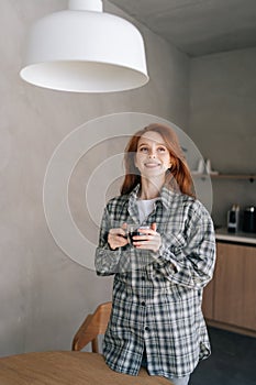 Vertical portrait of smiling woman enjoying holding cup drinking coffee standing in kitchen on sunny morning. Happy