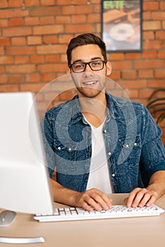 Vertical portrait of smiling young freelance programmer male in stylish glasses working on desktop computer sitting at