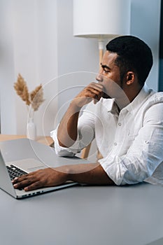 Vertical portrait of thoughtful black businessman working on business strategy on laptop sitting at desk, thinking