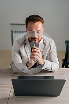 Vertical portrait of thoughtful businessman in suit holding smartphone sitting at office desk with laptop computer