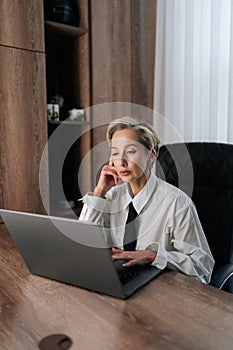 Vertical portrait of thoughtful middle-aged adult 50s woman working on laptop computer in office looking to screen