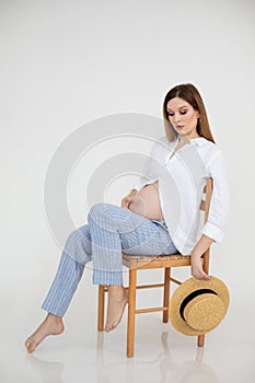 Vertical portrait of tired, fashionable barefoot woman with hat in hands, holding belly, looking at tummy. Resting pose