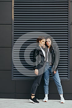 Vertical portrait of two girlfriends in casual clothes, wearing leather jackets and jeans, standing on a black background, posing
