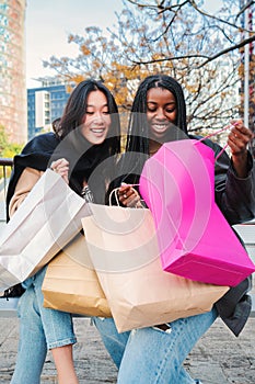 Vertical portrait of two multiracial young women or girls smiling and looking inside of a shopping bag sitting on a photo