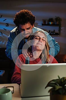 Vertical portrait of a woman tired and stressed from working with laptop, receives massage and caress from her husband