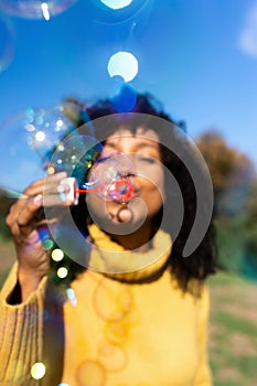 Vertical portrait of young african american woman making soap bubbles outdoors. Selective focus on bubbles.