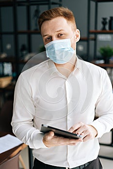 Vertical portrait of young business man wearing protective face mask using mobile phone standing in modern office room