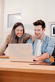Vertical portrait of young couple browsing on internet at home kitchen using a laptop. Happy husband and wife planning