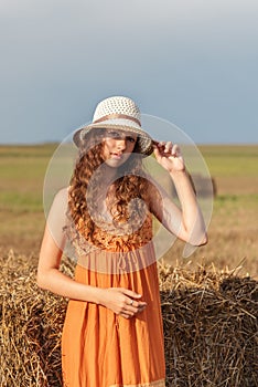 Vertical portrait of a young cute rural woman in an orange sarafan and a hat near a stack of wheat hay on a field in the rays of t
