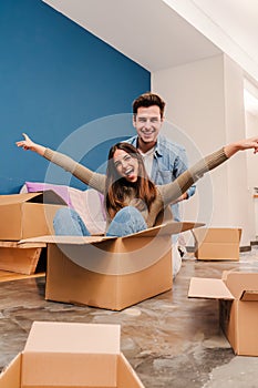 Vertical portrait of young happy couple having fun playing with cardboard box in their new home after moving. Husband