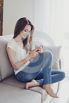 Vertical portrait of young pretty happy girl sitting on sofa using her pda. Girl is receiving and reading sms on her