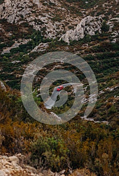 Vertical of a red car on the road through rocks covered with lush plants in the Massif des Calanques