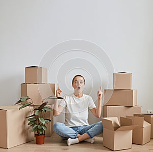Vertical shot of amazed woman wearing white T-shirt sitting on the floor near cardboard boxes with her belonging, posing during