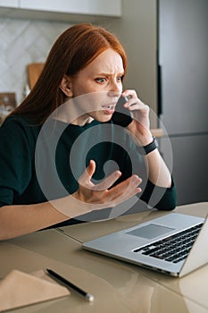 Vertical shot of angry young woman talking on mobile phone and using laptop sitting at table in kitchen with modern