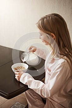 Vertical shot of attractive blonde daughter sitting in cozy pyjamas in kitchen, pouring milk in bowl with cereals