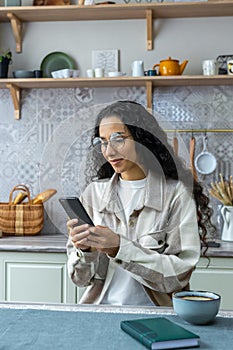 Vertical shot, beautiful hispanic woman using smartphone at home, woman with curly hair and glasses smiling, typing