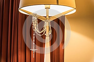 Vertical shot of a bedside lamp and a  silver cross hanging on it and shining under the lamp\'s light