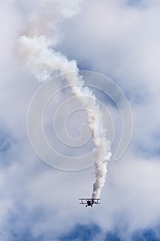 Vertical shot of a bi-plane stunt flying with a smoke trail in the blue sky with white clouds