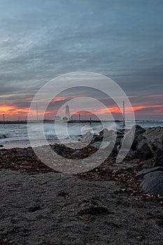 Vertical shot of big rocks at the beach with a seascape, lighthouse and sunset sky in the background