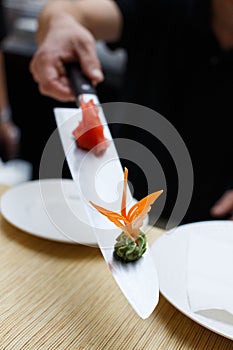 Vertical shot of a bokeh blurred male hand holding above a table a long kitchen knife with a small pile of red gari ginger and