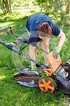 Vertical shot of brown haired female gardener raking fresh grass out lawn mower or grass cutter in garden. Agriculture