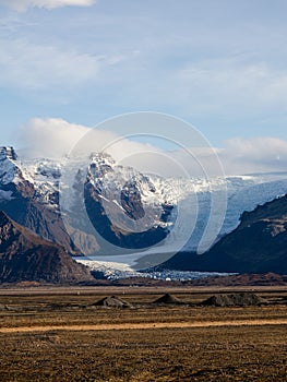 Vertical shot of a brown soil field with rocky mountains and glacier fading in from the sky, Iceland