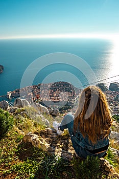 Vertical shot of a brunette girl sitting on top of the srd mountain looking down at the ancient Croatian city of Dubrovnik. Warm