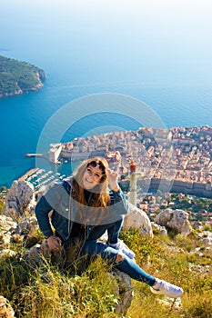 Vertical shot of a brunette sitting on the stones on the Srd mountain above the city of Dubrovnik, posing for a photo and laughing