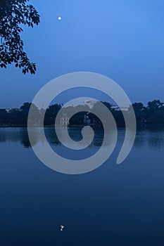 Vertical shot of a calm lake with the reflection of green trees and the full moon in the night sky