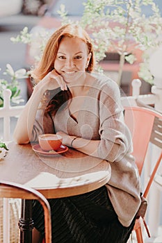 Vertical shot of cheerful foxy female dressed in fashionable clothes, drinks coffee, poses in outdoor cafe, enjoys spare time, has