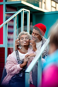 Vertical shot of cheerful man in trendy hat puts on headphones to his mulatto female friend, have entertainment together