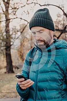 Vertical shot of concentrated hipster guy with thick beard, wears anorak and hat, holds cellular, uses special application for