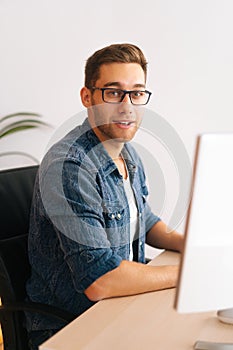 Vertical shot of confident young frelancer male in stylish glasses working on desktop computer sitting at desk at home