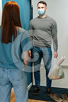 Vertical shot of courier male in protective face mask delivering groceries bag to female housewife customer on doorway