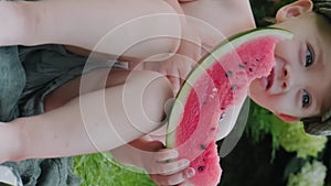 Vertical shot. Cute toddler boy sitting on grass eating watermelon outdoors.