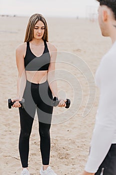 Vertical shot of a fit female doing bicep curls using dumbells with a male trainer at the beach