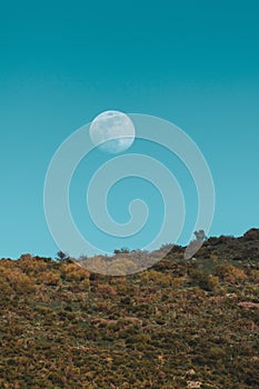 Vertical shot of a full moon on a blue sky above a hill at daytime