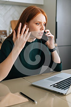 Vertical shot of furious young woman talking on mobile phone and using laptop sitting at table in kitchen with modern
