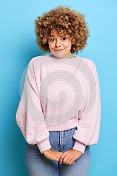 Vertical shot of gentle young woman with curly hair keeps arms together looks shy wears casual fluffy sweater and jeans