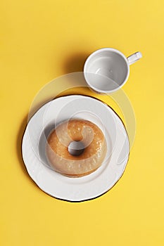 Vertical shot of glazed doughnuts in a white plate with a cup next to it on yellow background