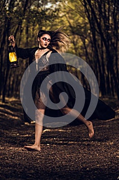 Vertical shot of a gothic girl in a transparent dress and black cape holding a lantern in forest