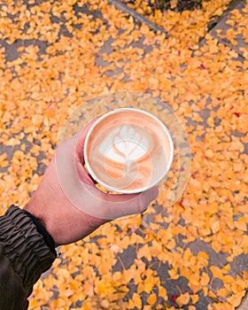 Vertical shot of a hand holding a white cup of latte with heart shape art foam against orange leaves