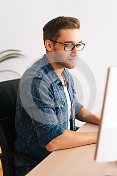 Vertical shot of handsome young designer male in glasses working on desktop computer sitting at desk at home office in