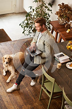 Vertical shot of handsome young man with a dog, leaning on table, holding laptop in hands, looking at screen, checking