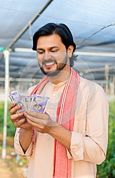 vertical shot of Happy smiling young farmer at greenhouse couting money or currency notes - concept of successful