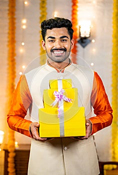 Vertical shot of happy smiling young indian man showing gift box by looking at camera during diwali celebration at home